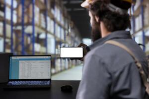 Storage room engineer using smartphone with white display, reading cargo details on distribution contracts. Supervisor with helmet looking at phone with blank mockup layout, warehouse logistics. photo