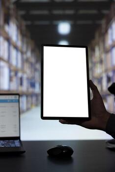 Storage room manager shows tablet with blank copyspace layout, working in a warehouse with racks filled of cargo in parcels and boxes. Engineer is looking at white screen on device display. photo