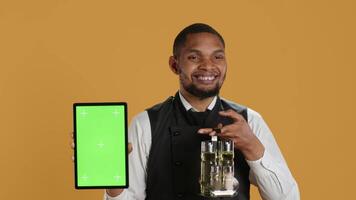 Professional waiter with apron presenting tablet with isolated mockup, presenting the copy space while he carries the cruet set. Skilled catering staff works in customer service at diner. Camera A. video