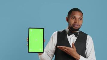Bellman hotelier showing a tablet with green screen display in studio, presenting mockup chroma key screen against blue background. Young hotel concierge works at resort. Camera A. video