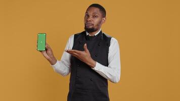 Restaurant staff showing a smartphone with green screen in studio, prepared to serve food orders in service oriented environment. Waiter with apron and uniform serving clients at tables. Camera B. video