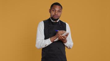 Skilled waiter with apron takes meal orders from clients on a notebook, writing down important information for customer satisfaction. Caterer butler working in service oriented industry. Camera B. video
