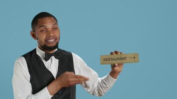 Doorman bellhop showing the restaurant direction with a hotel sign, holding an indicator to point guests to the amenities. Professional hotel concierge using a wall mark. Camera A. video