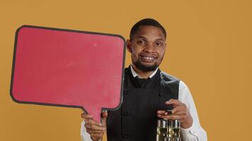 Catering employee showing a red speech bubble icon in studio, carrying a cruet set with condiments. Waiter with professionalism presenting an empty cardboard sign as an advertisement. Camera A. video