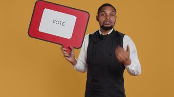 Professional waiter showing a message to vote on speech bubble, creating an advertisement for politics on cardboard icon. Restaurant employee working in customer oriented industry. Camera B. video