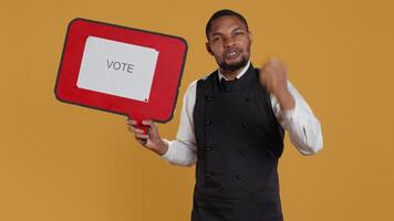 Waiter does an advertisement for politics and showing a speech bubble with the message to vote, standing against yellow background. Restaurant staff holds a cardboard sign. Camera B. video