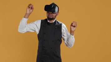 Restaurant waiter uses virtual reality headset with artificial intelligence, having fun with technology and vr glasses against yellow background. Employee with apron and uniform. Camera B. video