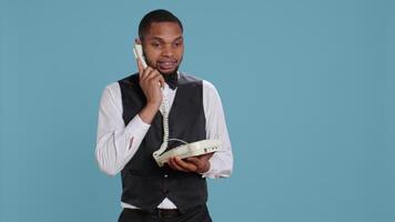 Bellboy concierge staff answering landline phone call to make reservations, discussing with clients about five star hotel rooms and amenities. Using telephone with cord to assist. Camera B. video
