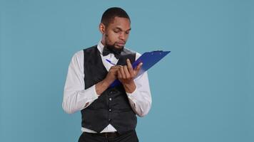 Receptionist worker writing down notes on clipboard files, ensuring easy check in process for hotel guests against blue background. Bellboy staff providing excellent concierge services. Camera B. video