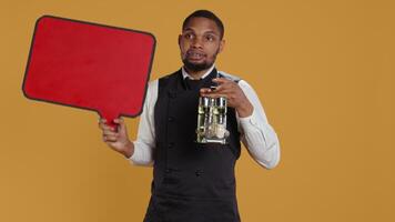 Catering employee showing a red speech bubble icon in studio, carrying a cruet set with condiments. Waiter with professionalism presenting an empty cardboard sign as an advertisement. Camera B. video