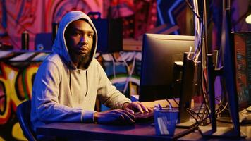 Portrait of african american hacker in abandoned warehouse using phishing technique that tricks users into revealing sensitive information. Hooded cybercriminal stealing passwords photo