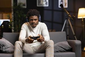 Gamer using headphones mic to discuss with teammates while playing games and enjoy leisure time. BIPOC man in apartment entertained by videogame on console, using gamepad photo