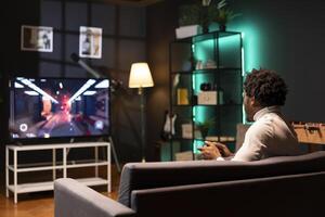 Man in home theatre using cloud gaming service to play demanding first person shooter videogame on TV. Gamer enjoying high quality FPS game, shooting enemies using gamepad photo