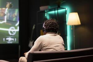 BIPOC man playing SF FPS videogame on widescreen smart TV, having fun in neon lit apartment. Gamer competing in online multiplayer game, using headphones to talk with teammates photo