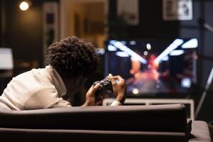 Man holding controller in apartment, playing videogames on smart TV display. Gamer on couch using joystick to participate in PvP online multiplayer game on console system attached to television set photo