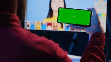 Photo editor using green screen mobile phone to watch tutorial, learning to color correct photographs in creative studio. Photographer watching guiding on chroma key smartphone