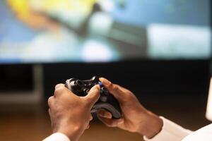 Close up shot of controller held by gamer sitting in front of large television set displaying videogame. Joystick used by person enjoying gaming session on widescreen smart TV at home photo