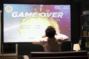 Gamer upset at seeing game over message on ultrawide TV screen playing arcade space shooter videogame. Man spending time at home on gaming system, frustrated after losing online competition photo