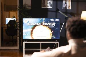 Man playing videogames on smart TV display, receiving game over screen on gaming console. Gamer upset after getting his spaceship exploded by rival players in online exports competition photo