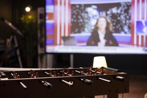 Focus on tabletop football game in front of ultrawide smart TV in blurry background displaying news program broadcasting. Big television screen in empty room showing VOD channel with newscast coverage photo