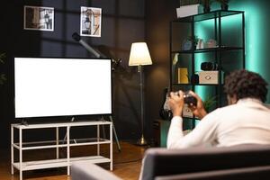 Gamer at home using mockup display television set to do gaming with controller. Man sitting on couch in living room, playing videogame on isolated screen smart TV to take off steam photo