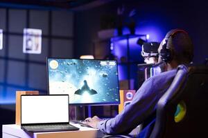 Isolated screen laptop next to professional streamer competing in online multiplayer videogame, streaming live at home. Mockup notebook and man playing on PC, using headphones to talk with audience photo