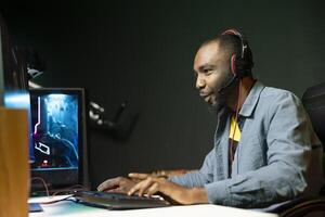 Smiling gamer with headphones on relaxing in gaming room, playing videogames on powerful computer. Cheerful esports player performing in gaming tournament from home late at night photo