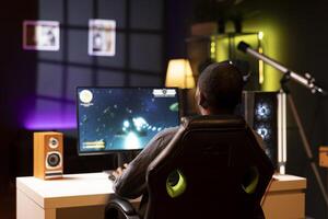 African american man playing SF space videogame on PC screen in neon illuminated apartment living room. Gamer enjoying spaceship flying game on computer desktop display in RGB lit home late at night photo