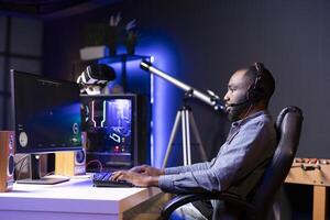 Man in RGB lit apartment playing immersive games on desktop PC at computer desk, unwinding after work. African american gamer competing against enemies in internet multiplayer shooter photo
