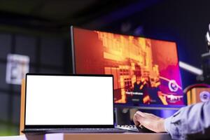 Mockup laptop display next to player using keyboard to play FPS videogame, shooting enemies. Isolated screen notebook near person gaming on computer desktop at home, close up shot photo