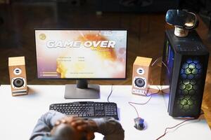 Heartbroken gamer dejected after losing in online multiplayer videogame, being defeated by rivals. Man putting hands on head in frustration after seeing game over screen on gaming pc monitor photo