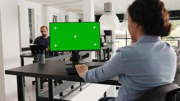 Employee sitting at desk with greenscreen on monitor, looking at isolated copyspace layout on computer desktop in coworking space. Businesswoman working on pc, blank mockup template. photo