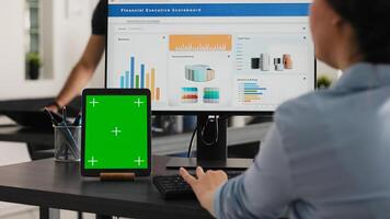 Specialist works with greenscreen tablet in open floor plan office, presenting isolated display on modern device. Businesswoman using pc monitor and blank chromakey layout on mini screen. photo