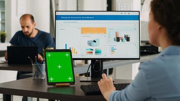 Agency employee looking at tablet with greenscreen while she is typing information on computer, checking mockup template on modern gadget. Woman working on business with chromakey in office. photo