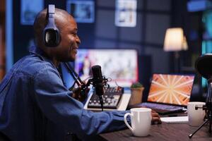 Smiling black man hosting a talk show is shown in a side view profile, talking with his online listeners while doing a live podcast recording from his home studio, he is sporting wireless headphones. photo