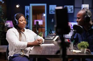 Influential African American host engages with guest, using quality podcast equipment in their stylish home studio. Black couple utilizing smartphone on tripod to record their online talk show. photo