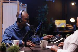 African American male radio host using professional equipment for live broadcasting, featuring an audio mixer and microphone setup. Black man preparing sound gadgets for efficient podcast streaming. photo