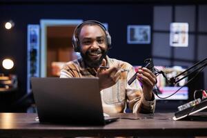 Cheerful tech expert going on-air, describing details of new mobile device for online viewers. Smiling black man using professional recording equipment to showcase newly released cell phone. photo