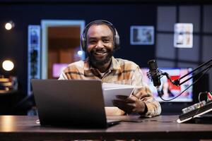 Cheerful black guy presents variety of things to online audience while carrying a notepad and gazing into the camera. Joyful African nfluencer reads a script from a book while creating content. photo