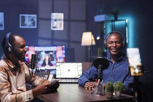 Cheerful vloggers streaming podcast using smartphone on tripod during live debate on vlog channel. Excited men meeting in studio to film show with mobile phone and talk global events. photo