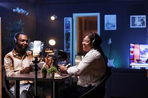 Journalist records an interview with phone on tripod stabilizer in studio, reviewing notes to ask questions to guest. Black man and woman using mobile device for vlogging at home. photo