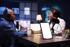 Two African American vloggers wearing headphones are recording a live podcast while a tablet on desk shows a white screen with mockup template. Digital device on table displays isolated chromakey. photo