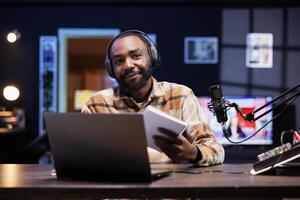 Black male vlogger holding a notebook and looking at camera, presenting various topics for his online viewers. African american influencer records content, reading a script from a book. photo