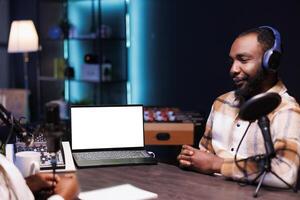African man wears headset and speaks into mic while his laptop displays blank white screen. Device on desk shows an isolated chromakey mockup template while male content creator making a podcast. photo