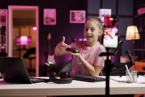 Cute girl in studio reviewing neon lit gaming mouse in front of camera in personal studio. Joyous kid filming electronics haul, presenting Bluetooth wireless computer peripheral to viewers photo