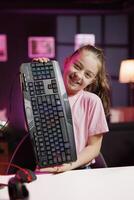 Happy gen Z content creator filming keyboard review in home studio, showing typing capabilities. Smiling small girl showcasing RGB illuminated computer peripheral to audience photo