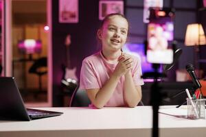 Joyful small girl shooting for social media platform using smartphone. Young media star entertaining followers, doing content creation in pink neon lit apartment using phone on tripod photo