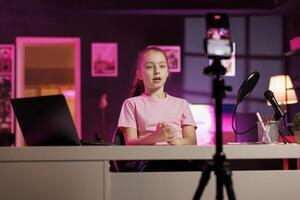 Child doing content creation, using cellphone on tripod stabilizer to film vlog channel intro. Young online star greets children audience, presenting today topic, recording with mobile phone photo