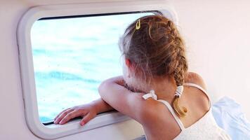 Blond little girl sits in the cabin of a yacht and looks out the window during a sea crossing. Yacht life concept. High quality 4k footage video
