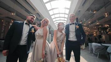 The groomsmen and bridesmaids are captured smiling and enjoying themselves at an elegant wedding venue. The bridesmaids wear matching light-colored dresses, and the groomsmen are dressed in sharp video